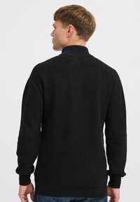 Svart ribbestrikket genser med høy krage og lange ermer. Har teksturert stoff, tettsittende mansjetter og en rett kant bak.