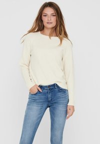 Helle Creme Strickpullover mit rundem Halsausschnitt, kombiniert mit eng anliegenden blauen Jeans im klassischen Fünf-Taschen-Design und leichter Ausbleichung.