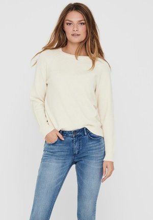 Lys kremfarget strikkegenser med rund hals, kombinert med tettsittende blå jeans med klassisk fem-lomme-design og lett falming.