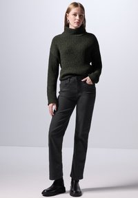 Maglione lavorato a maglia verde scuro con colletto alto, abbinato a pantaloni neri a gamba dritta e stivaletti neri, esposto su uno sfondo chiaro.