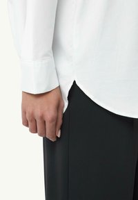 Biała bluzka z długim rękawem i mankietami na guziki, z zaokrąglonym dołem, w zestawieniu z czarnymi spodniami. Materiał wydaje się gładki i sztywny.