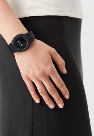 Black sporty watch with a rubber strap, round face, and distinct hour markers; worn on a hand resting against a black garment.