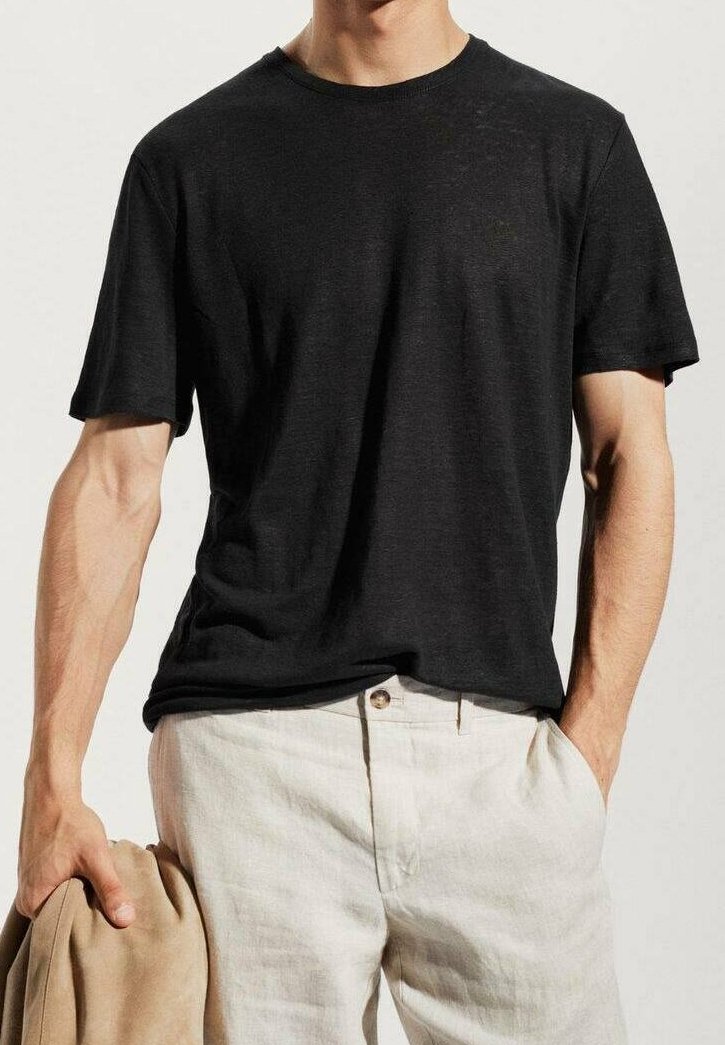 T-shirt noir à manches courtes en tissu doux, col rond, coupe décontractée, porté avec un pantalon en lin beige clair, une main tenant une veste beige.