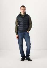 Black puffer vest with zip closure over a green hoodie, paired with blue jeans and black shoes. Simple design, flat texture, no visible patterns.