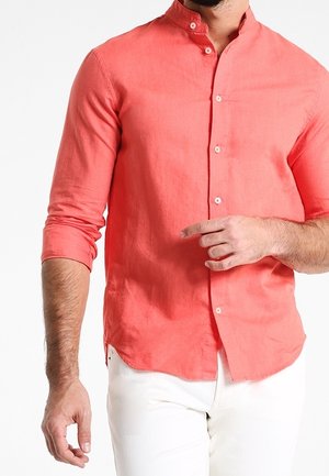 Mężczyzna w koralowo-czerwonej koszuli z długim rękawem i kołnierzem mandaryńskim, z podwiniętymi rękawami i białych spodniach na białym tle.