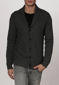 Grå stickad cardigan med sjalkrage, sex knappar, ribbade mudd och nederkant; texturerat mönster på kroppen, bärs över en svart skjorta.