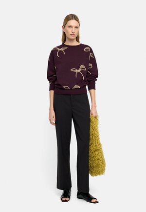 Kobieta w ciemnofioletowym swetrze z beżowymi wzorami kokard, czarnych spodniach, czarnych botkach z odkrytymi palcami, trzyma dużą, puszystą żółtą torbę.