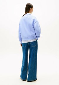 Felpa lilla con orlo a costine, indossata sopra una camicia a righe bianco e blu. Abbinata a jeans blu a gamba larga.