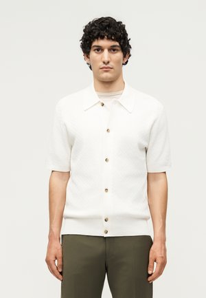 Homme aux cheveux courts, bouclés et noirs, portant une chemise blanche boutonnée à manches courtes et un pantalon vert olive, debout devant un fond clair uni.