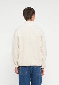 Kreemivärvi fliisist pullover kõrge kraega, langetatud õlgadega ja soonikute otstega. Omab kontrastset musta äärt ja tekstureeritud kangast.
