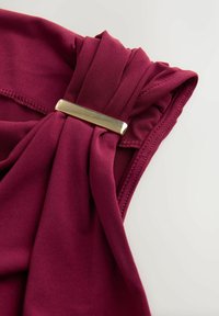 Burgunderfarbener Stoff mit gefaltetem Design und einem goldenen Metallakzent. Glatte Textur mit geschwungenen Nähten entlang der Kanten.