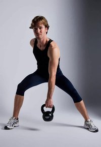 Fekete kettlebell tartása guggolásban, egy fiatal férfi viseli, fekete ujjatlan pólóban és sötétkék kompressziós leggingsben. Világosszürke háttér.