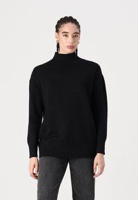 Fekete kötött garbó pulóver bordázott mandzsettákkal és aljjal. Laza fazon; sima текстура; réteges kialakítás a plusz melegség érdekében. Sötétszürke nadrággal párosítva.