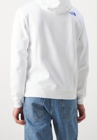 Bílá mikina s kapucí a žebrovanými manžetami a spodním lemem, s modrým logem značky The North Face na horní části zad. Nosit s modrými džíny.