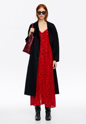 Kobieta w czarnym płaszczu na czerwonej sukience w czarne kropki, czarnych butach, ciemnych okularach przeciwsłonecznych, z ciemnoczerwoną torebką na ramieniu.