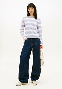Top de manga larga a rayas en rosa claro y lavanda, combinado con jeans oscuros holgados y zapatillas crema, sosteniendo un bolso blanco suave.