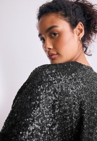 Haut noir à sequins avec une coupe ample, présentant des sequins brillants et réfléchissants en surface ainsi qu'une texture douce. Vue de dos mettant en valeur des manches surdimensionnées.