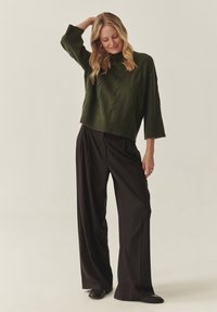 Tumši zaļš adīts džemperis ar saīsinātu dizainu, kombinēts ar melnām platajām biksēm un plakaniem apaviem. Ir redzama teksturēta adījuma detaļa.