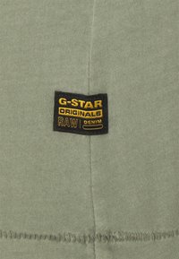 Grön tyg med en synlig svart etikett som står "G-STAR ORIGINALS RAW DENIM" i gult, sydd på plagget. Slät textur, närbild.