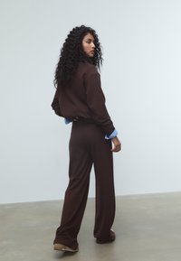 Ruskea neulepaita, jossa on rento istuvuus ja siniset hihansuut, yhdistettynä leveälahkeisiin ruskeisiin housuihin ja ruskeisiin kenkiin, vaaleaa taustaa vasten.