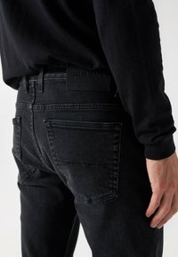 Črne denim hlače z elegantnim dizajnom, vidnim šivom in dvema zadnjima žepoma. Oznaka logotipa nosi napis "SALSA JEANS" in je nameščena na pasu.