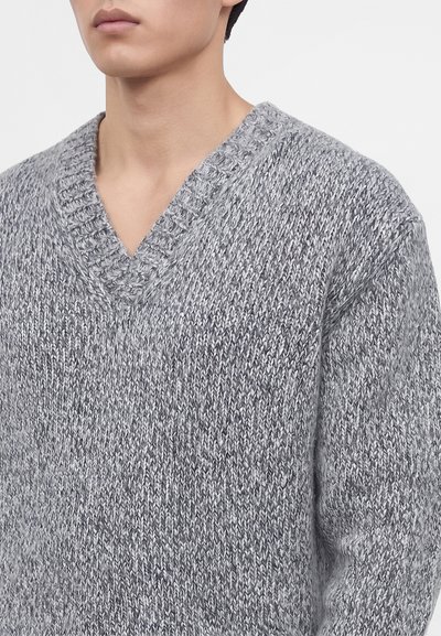 Personne portant un pull en maille gris chiné à col en V avec un col côtelé texturé sur un fond clair uni.