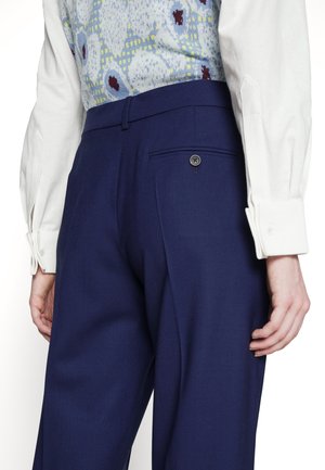 Person trägt dunkelblaue High-Waist-Hose mit Knopftasche hinten, weißes langärmeliges Hemd und eine gemusterte blaue Weste.