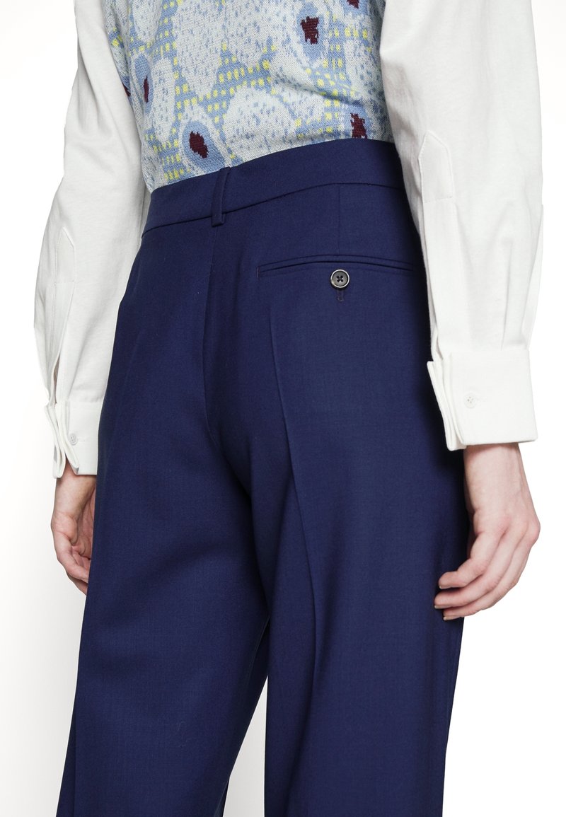 Personne portant un pantalon taille haute bleu foncé avec une poche arrière à bouton, une chemise blanche à manches longues et un gilet bleu à motifs.