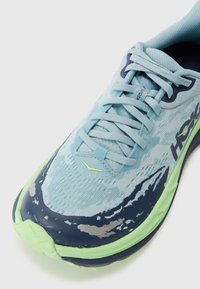 Baskets de course bleu clair avec un dessus en mesh texturé, accents bleu foncé et vert, et détails gris sur le bout et la semelle.