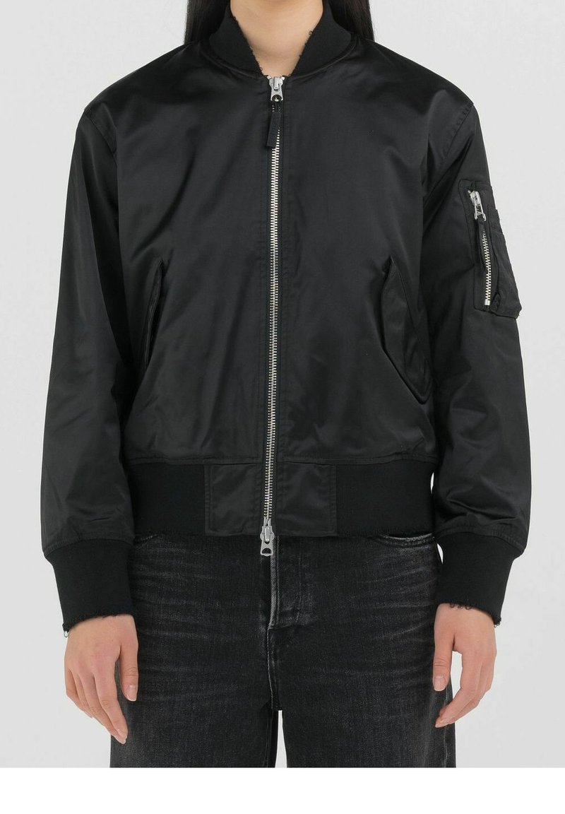 Personne portant une veste bomber noire avec des poches zippées à l'avant et sur les manches, associée à un jean noir sur un fond clair uni.