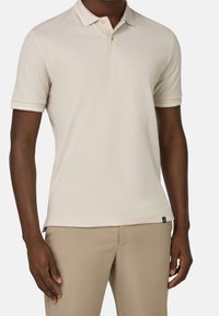 Beige pikétröja med en tvåknappslå, kort ärm och kontrasterande krage. Slät textur och minimal logotyp vid fållen.