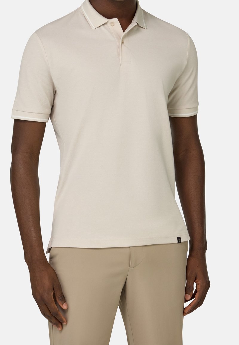 Beige pikétröja med en tvåknappslå, kort ärm och kontrasterande krage. Slät textur och minimal logotyp vid fållen.