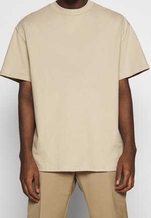 Person wearing a plain beige short-sleeve t-shirt and matching beige pants, standing with arms relaxed at their sides.