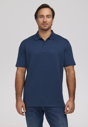 Mężczyzna w teksturowanej granatowej koszulce polo i niebieskich dżinsach, stojący na jednolitym jasnoszarym tle.