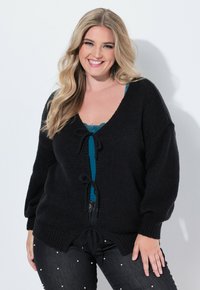 Svart stickad kofta med V-ringning, lös passform och knytningar, bärs över en tealfärgad spetscamisole, i kombination med mörk denim dekorerad med pärlor.