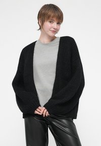Cardigan nero a maglia con una vestibilità ampia, maniche larghe e scollo a V profondo, abbinato a una maglietta grigia e pantaloni in pelle nera lucida.