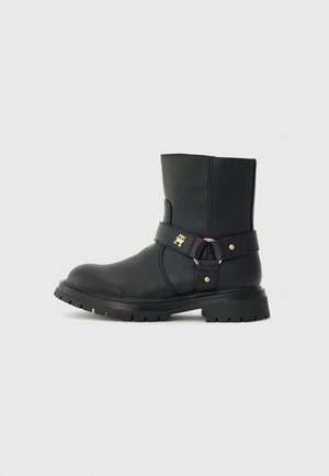 Bottines en cuir noires mi-mollet avec semelle épaisse, sangle décorative, boucle en or et petit logo doré sur le côté, sur fond uni.