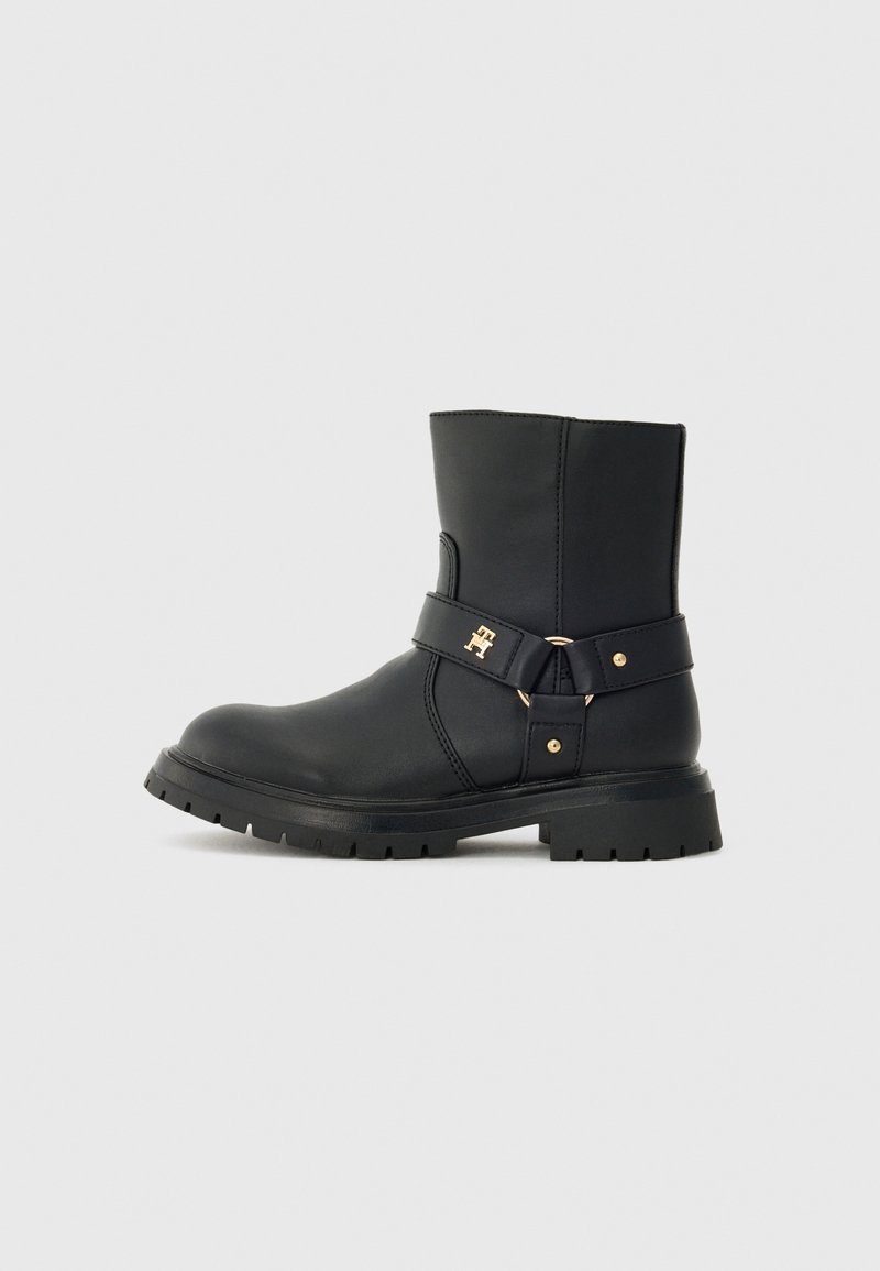 Bota de couro preta até ao meio da panturrilha com sola grossa, faixa decorativa, fivela dourada e pequeno logotipo dourado na lateral, contra um fundo simples.