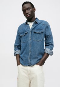 Chemise en denim bleu à boutons, dotée de poches poitrine doubles, de manches retroussées et d'une texture douce. Associée à un pantalon de couleur crème.