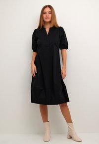 Svart klänning med knälång fåll, puffärmar och knappslå. Modellen kombinerar den med ljusa ankelkängor. Slät tygtextur.