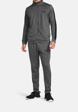 Man draagt een donkergrijs Under Armour trainingspak met zwarte strepen langs de mouwen en broekspijpen, gecombineerd met zwarte sportschoenen.