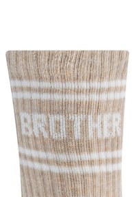 Beżowa skarpetka dzianinowa z białymi paskami i napisem "BRAT" napisanym dużymi literami, prezentująca teksturowany materiał i cylindryczny kształt.