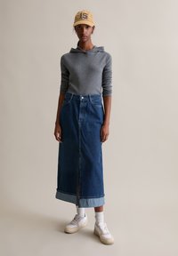 Grijze hoodie met lange mouwen, high-waisted blauwe denim maxi-rok met een voorwerp en witte sneakers met beige accenten. Pet met nummer 15.