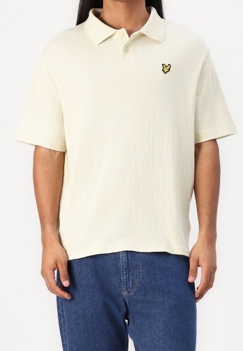 Polo à manches courtes de couleur jaune clair, en tissu texturé, avec un col et un petit logo noir sur la poitrine. Porté avec un jean bleu.