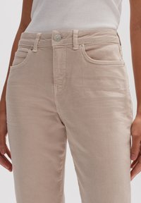 Jean beige en mélange de coton avec une texture lisse, cinq poches, fermeture par bouton argenté et détails de couture subtils. Coupe ajustée à jambe droite.