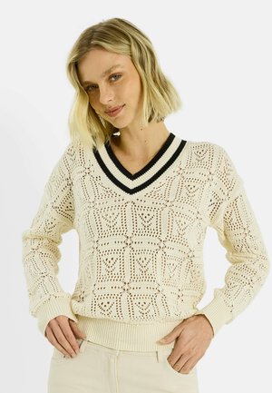 Mujer rubia con un suéter de crochet color crema con escote en V y ribete negro, posando con las manos en las caderas sobre un fondo blanco liso.