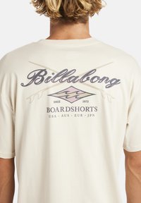 Achteraanzicht van een persoon in een crèmekleurig Billabong T-shirt met gekruiste surfplanken en de tekst "Boardsports USA AUS EUR JPN" op de achterkant.