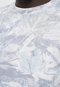 Γκρι μπλουζάκι με διακριτικό σχέδιο φύλλων, κατασκευασμένο από απαλό ύφασμα με στρογγυλή λαιμόκοψη και ραφές χωρίς ραφές στους ώμους.