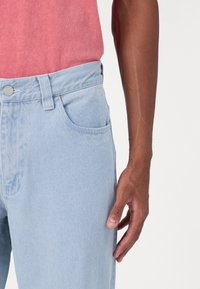 Ανοιχτού μπλε τζιν παντελόνια με κλασικό σχέδιο πέντε τσέπες, κλείσιμο με μεταλλικό κουμπί και διακριτική υφή. Χέρι που ακουμπάει στο πλάι.