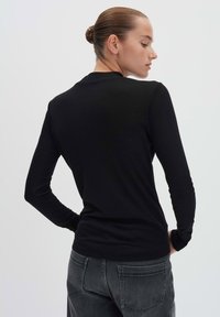Vrouw met een knot in haar haar, gekleed in een zwart shirt met lange mouwen en donkergrijze jeans, staand met haar handen op haar heupen, zijwaarts kijkend tegen een witte achtergrond.